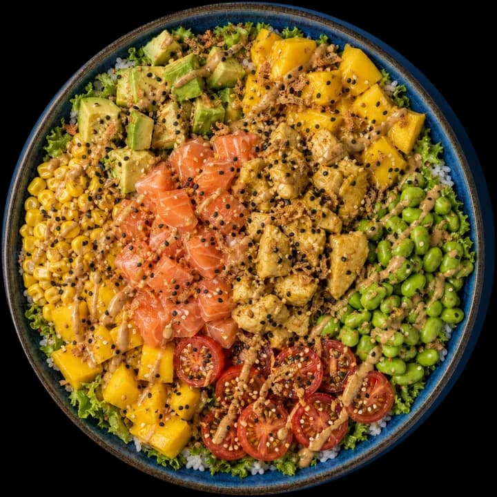 Exemple de bowl en taille L, vu de dessus : riz à sushi + batavia verte, avocat, edamame, mangue ×2, tomates cerises, maïs, saumon cru, poulet mariné, sésame grillé, oignons frits, cacahuète