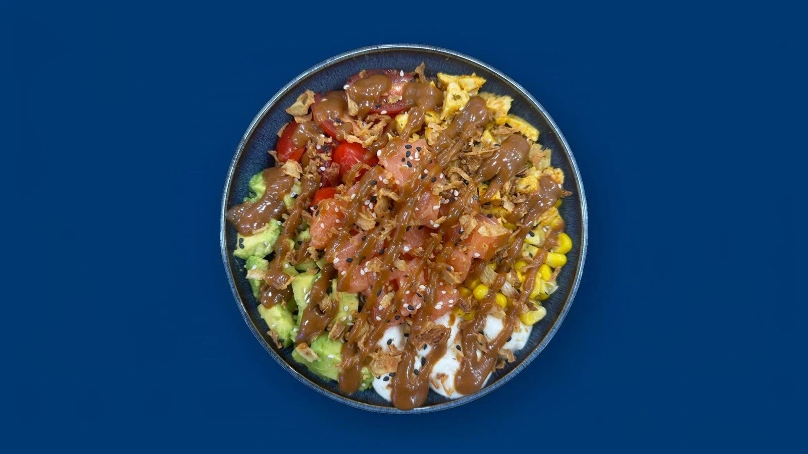 Le bol composé, vu de dessus : batavia, riz à sushi, tomates cerises, avocat, mozzarella, maïs, poulet mariné, saumon cru, sésame grillé, oignons frits et sauce cacahuète